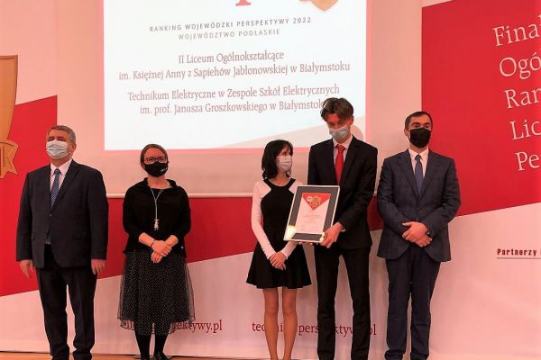 Zdjęcie z dyplomem na tle ogłoszonych wyników